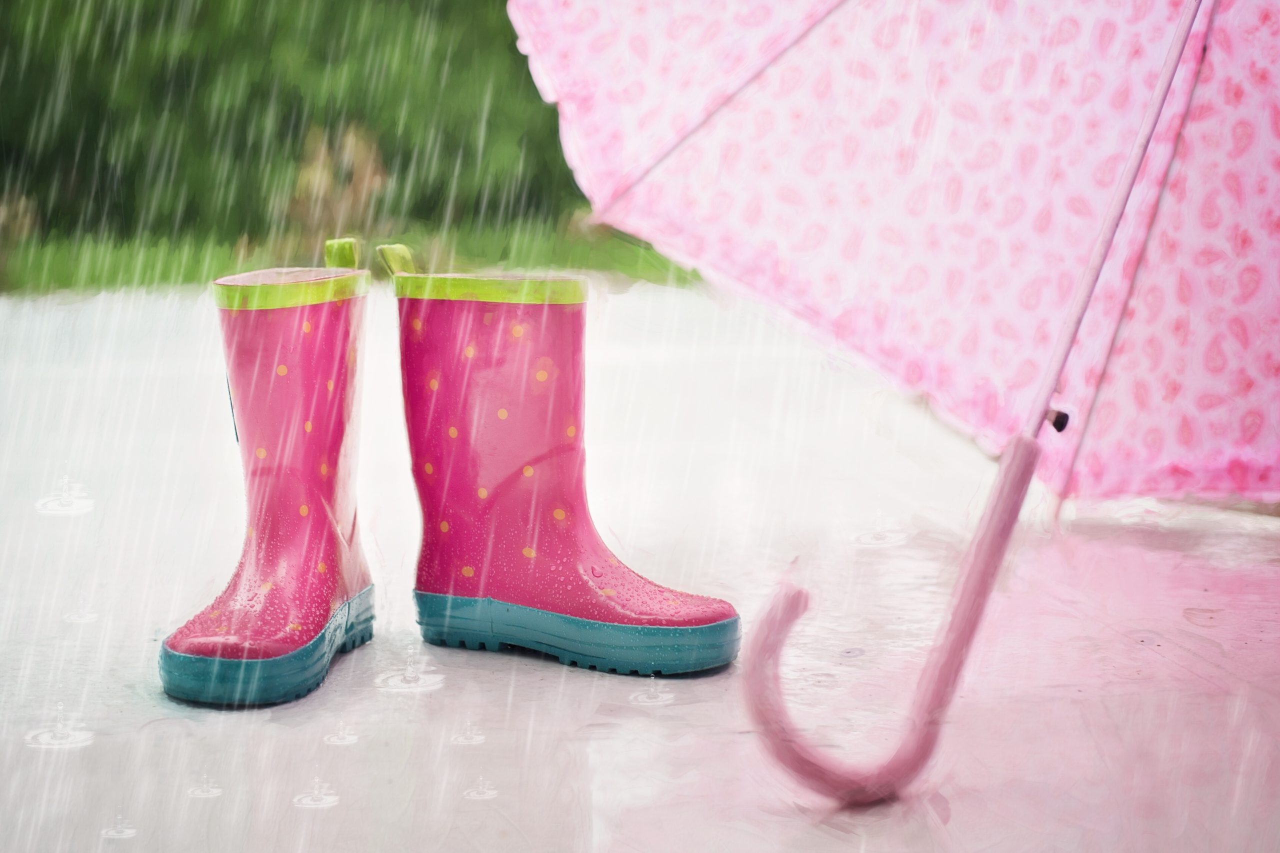 a pair of pink rainboots next to a pink umbrella. Rain is falling in the background. This image represents spring time. 